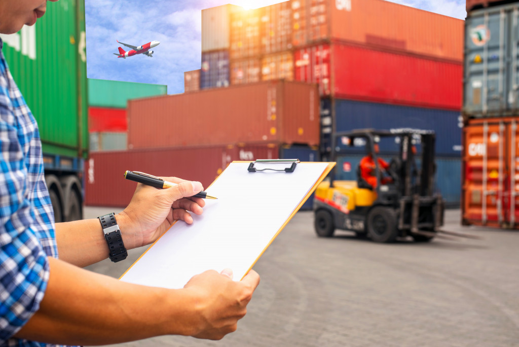 a man holding a clipboard and auditing while facing container boxes and a forklift
